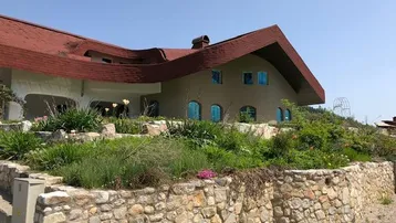 Auffälliges Haus mit einzigartigem Dach und einer Natursteinmauer im Vordergrund, umgeben von Grün.