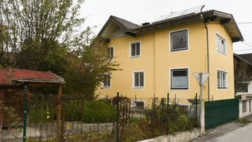 Gelbes Wohnhaus mit Satteldach und Photovoltaikanlage, umgeben von Bäumen und einem Zaun.