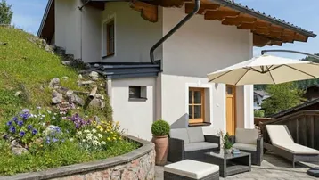 Gemütliche Terrasse mit Gartenmöbeln, Sonnenschirm und blühenden Blumenbeeten am Hang.