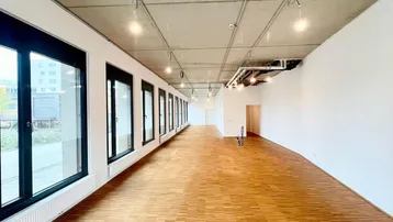 Langer, heller Gewerberaum mit großen Fenstern, Holzboden und Sichtbetondecke, bereit zur individuellen Gestaltung.