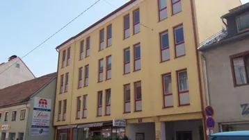 Gelbes Mehrfamilienhaus mit mehreren Stockwerken und Geschäften im Erdgeschoss an einer Straße.