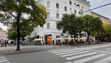 Weißes Eckgebäude mit klassischer Architektur an einer belebten Straße mit Bäumen und Fußgängerüberweg.