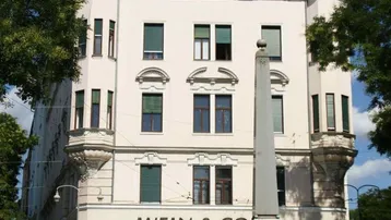 Historisches Gebäude mit markanter Architektur und Geschäft im Erdgeschoss, umgeben von Bäumen und einem Denkmal.