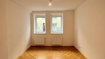 Heller, leerer Raum mit Parkettboden und zwei Fenstern, ideal als Wohnzimmer.