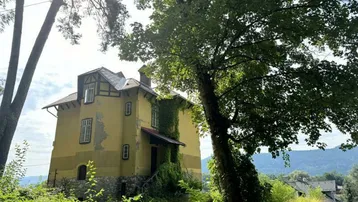 Historisches Gebäude mit gelber Fassade, umgeben von altem Baumbestand und üppigem Grün.