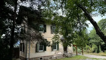 Historisches Gebäude mit verwitterter Fassade, umgeben von altem Baumbestand und viel Grün.