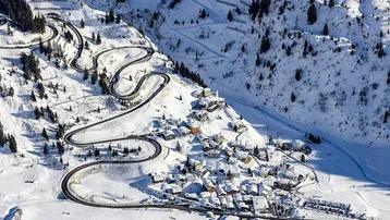 Luftaufnahme eines verschneiten Bergdorfes mit Serpentinenstraße und umliegenden Wäldern im Winter.