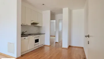 Kleine Einbauküche mit weißen Schränken, Holzparkettboden und grundlegenden Geräten in offenem Raum.