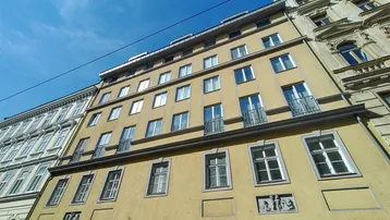 Fassade eines Mehrfamilienhauses mit gelbem Putz und kleinen Balkonen unter blauem Himmel.