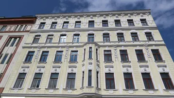 Historische Fassade eines Mehrfamilienhauses mit vielen Fenstern, Erker und dekorativen Elementen unter blauem Himmel.