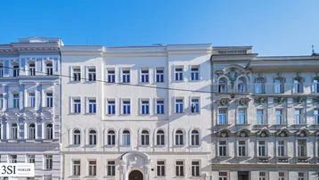 Die helle Fassade eines Altbaus mit vielen Fenstern unter blauem Himmel.
