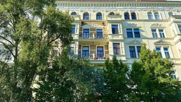 Historische Fassade eines Mehrfamilienhauses mit verzierten Balkonen und grünen Bäumen davor.