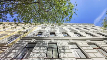 Historische Fassade eines Altbaus mit vielen Fenstern und grünen Bäumen im Vordergrund.