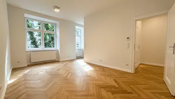Helles Zimmer mit Fischgrätparkettboden und großen Fenstern, das viel Tageslicht hereinlässt.