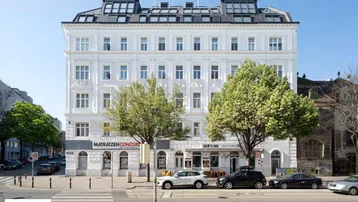 Weißes Mehrfamilienhaus mit klassischer Fassade, Geschäften im Erdgeschoss und Bäumen davor.
