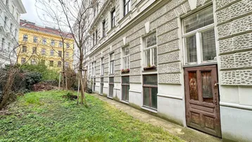 Seitenansicht eines Altbaus mit Fenstern und einer Holztür, angrenzend an eine begrünte Fläche mit Bäumen.
