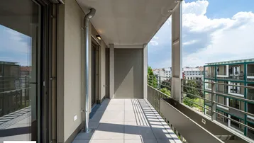 Langer, schmaler Balkon mit Fliesenboden und Blick auf umliegende Gebäude unter blauem Himmel.