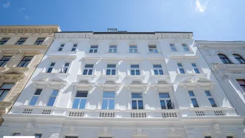 Weiße Fassade eines historischen Gebäudes mit vielen Fenstern und dekorativen Elementen unter blauem Himmel.