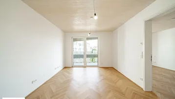 Helles, leeres Zimmer mit Fischgrätparkett und Zugang zu einem Balkon in einem Neubau.
