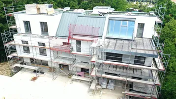 Im Bau befindliches Mehrfamilienhaus mit Gerüst, umgeben von Bäumen und Hügeln.