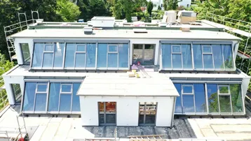Gebäude im Bauzustand mit moderner Dachkonstruktion und großen Glasflächen, umgeben von Bäumen.