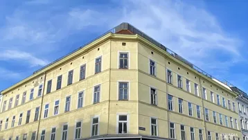 Eckgebäude mit gelber Fassade, vielen Fenstern und einem Dachterrassenbereich unter blauem Himmel.