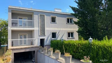 Graues Mehrfamilienhaus mit zwei Balkonen und einer Garageneinfahrt, umgeben von grünen Hecken.