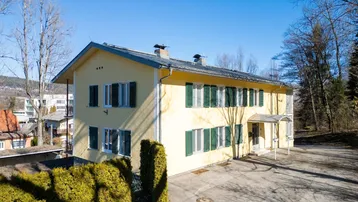 Gelbes Mehrfamilienhaus mit grünen Fensterläden und großem Vorplatz unter blauem Himmel.