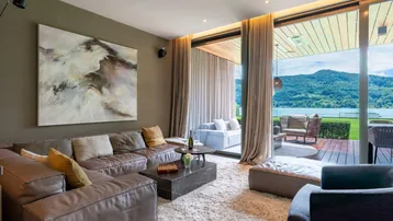 Geräumiges Wohnzimmer mit bequemer Ledercouch, modernem Kunstwerk und direktem Zugang zur Terrasse mit Seeblick.