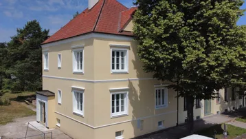 Klassisches Gebäude mit gelber Fassade, weißen Fenstern und rotem Satteldach unter blauem Himmel.
