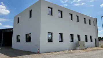 Modernes, hellgraues Gebäude mit vielen Fenstern unter einem blauen Himmel und ländlicher Umgebung.