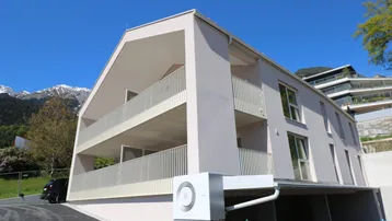 Modernes Mehrfamilienhaus mit Balkonen und Wärmepumpe vor alpiner Bergkulisse.
