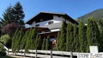 Hausfassade im alpinen Stil mit gepflegter Hecke und Bergblick unter blauem Himmel.