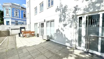 Eine gepflasterte Terrasse mit Holzbänken und Tischen vor einem weißen Gebäude, im Hintergrund ein blaues Haus.