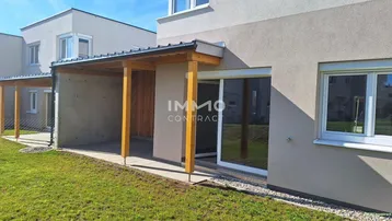 Überdachte Terrasse mit Holzbalken und angrenzendem Rasenbereich eines Neubaus.