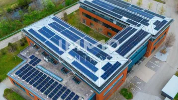 Luftaufnahme eines modernen Bürogebäudes mit zahlreichen Solarmodulen auf dem Dach, umgeben von grünen Feldern und einem Fluss.