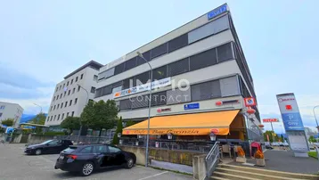 Großes Geschäftsgebäude mit Café im Erdgeschoss und Parkplätzen davor unter bewölktem Himmel.