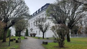 Großes, gut erhaltenes Gebäude mit Mansarddach, umgeben von einem parkähnlichen Garten mit alten Bäumen.