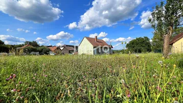 Ländliches Haus umgeben von einer weiten Wiese unter blauem Himmel mit Wolken.