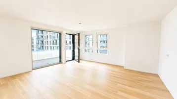 Helles, leeres Wohnzimmer mit Parkettboden, großen Fenstern und Zugang zum Balkon.