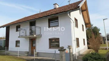Einfamilienhaus mit weißer Fassade, Satteldach und kleinem Balkon im Vorgartenbereich.