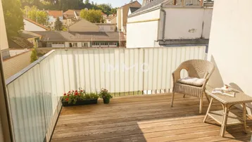 Sonniger Balkon mit Holzboden, Sitzgelegenheiten und Blick auf die umliegenden Häuser.
