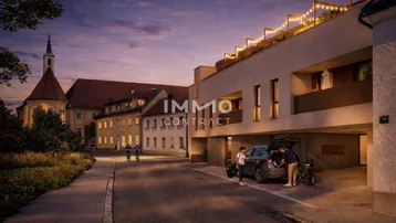 Abendliche Außenansicht eines modernen Wohngebäudes mit beleuchteter Terrasse und historischer Kirche im Hintergrund.