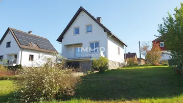 Weißes Einfamilienhaus mit Satteldach und Balkon, umgeben von einer grünen Wiese.