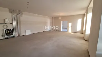 Großer, leerer Raum im Rohbauzustand mit Betonboden und sichtbaren Installationen, durchflutet von natürlichem Licht.