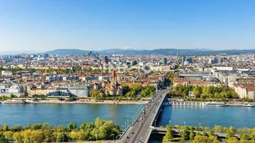 Panoramablick über eine Stadt mit Fluss, Brücke und Bergen im Hintergrund unter blauem Himmel.