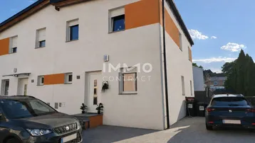 Modernes Reihenhaus mit weißer Fassade, orangefarbenen Akzenten und zwei Parkplätzen.