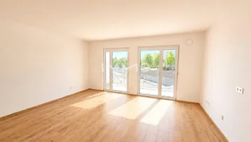 Helles, leeres Zimmer mit Holzboden und großen Fenstertüren, die zu einem Außenbereich führen.