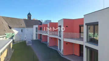 Reihenhäuser in moderner Architektur mit leuchtend roter Fassade, Balkonen und Blick auf eine Grünfläche.