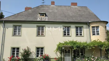 Historisches Gebäude mit gepflegtem Vorgarten, Rosenbüschen und traditioneller Architektur unter blauem Himmel.